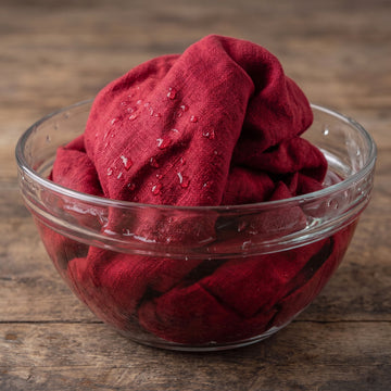 A deep red textured linen fabric cloth,  filled with water droplets and presented inside a clear glass bowl on a rustic wooden surface.