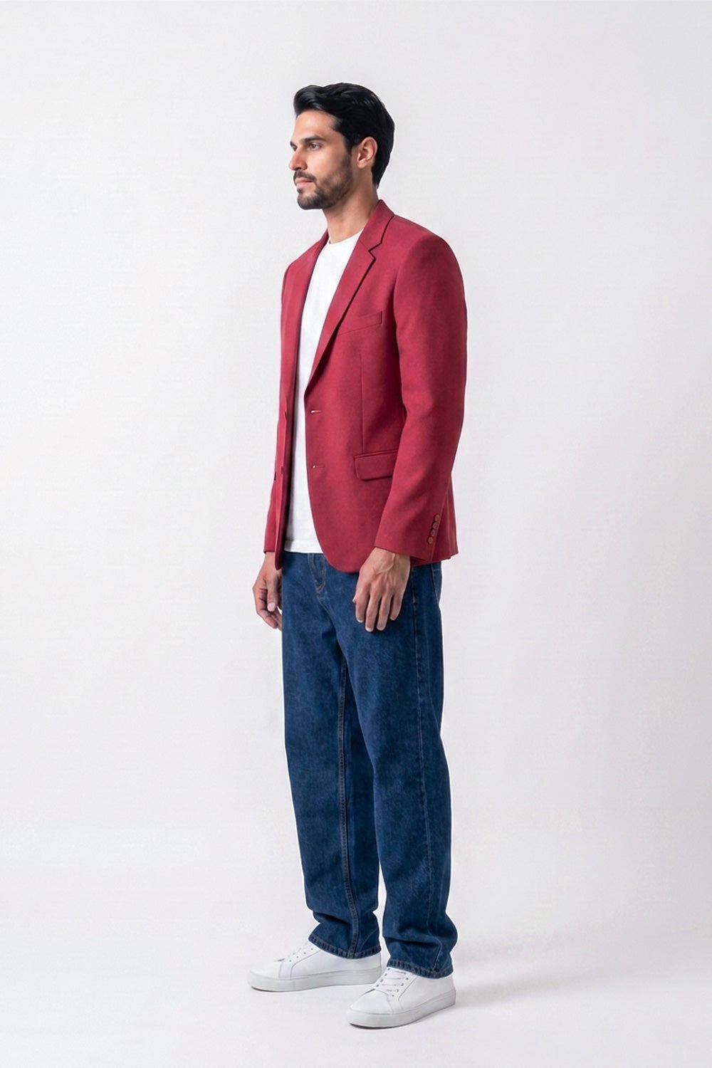 A man wearing a red linen blazer over a white t-shirt, blue jeans, and white sneakers, standing in profile against a plain white studio background.