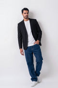 A man wearing a black linen blazer, white t-shirt, blue jeans, and white sneakers, standing against a plain light gray studio background.