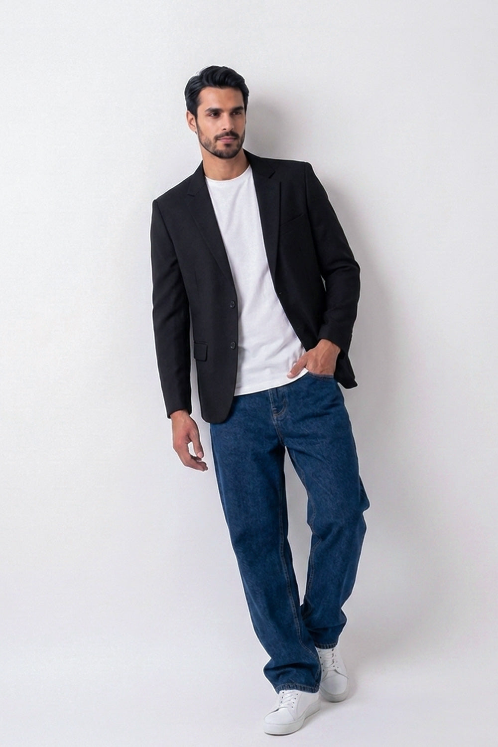 A man wearing a black linen blazer, white t-shirt, blue jeans, and white sneakers, standing against a plain light gray studio background.