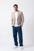 A man modeling a beige linen blazer over a white t-shirt, paired with dark blue jeans and white sneakers, standing against a plain white studio background.