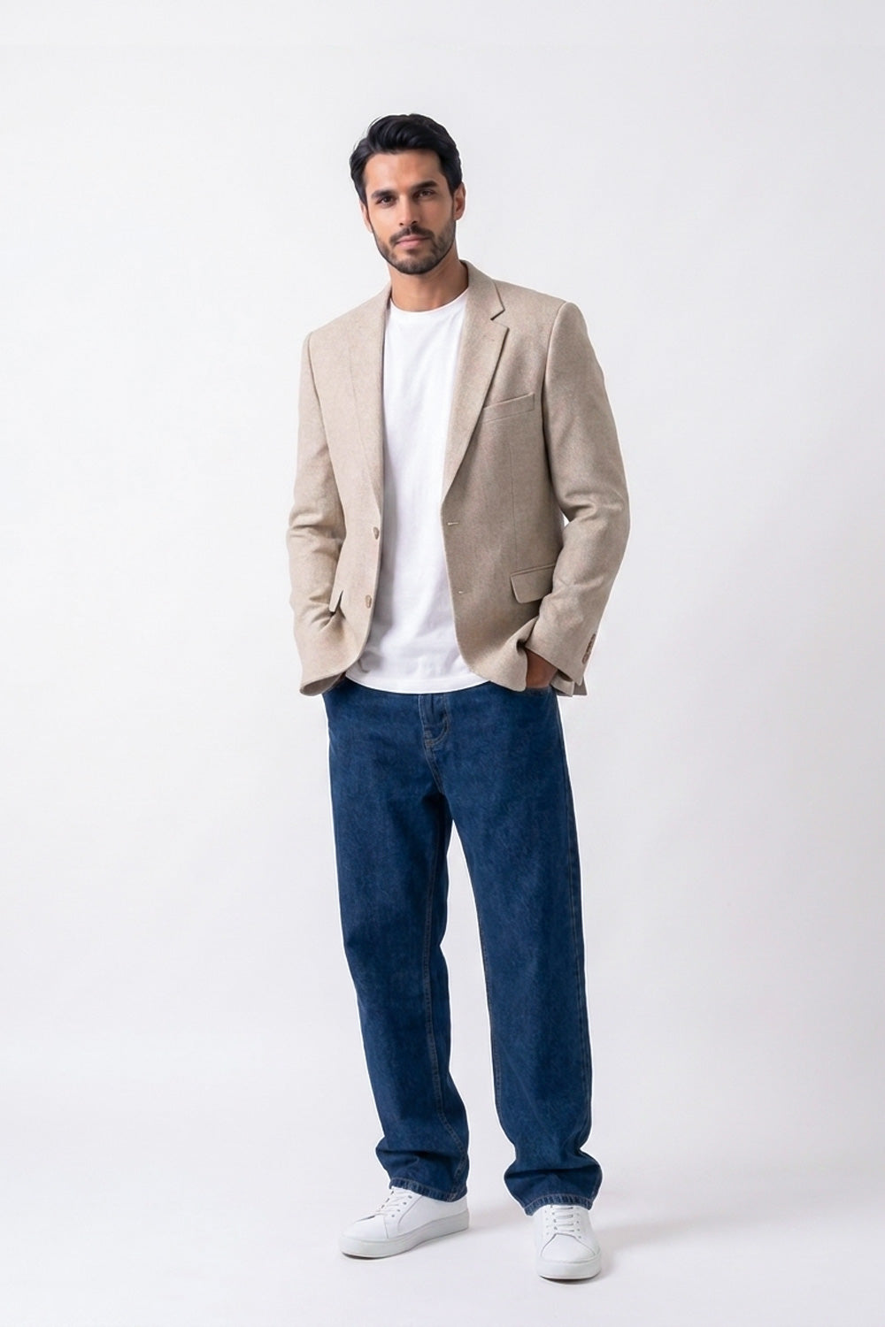 A man modeling a beige linen blazer over a white t-shirt, paired with dark blue jeans and white sneakers, standing against a plain white studio background.