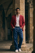 A man stands in a stone corridor wearing a red linen blazer, white dress shirt, blue jeans, and white sneakers.