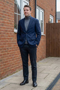 A man wearing a navy blue linen blazer, dark trousers, a gray turtleneck, and black dress shoes, standing outdoors on a paved walkway next to a red brick wall.