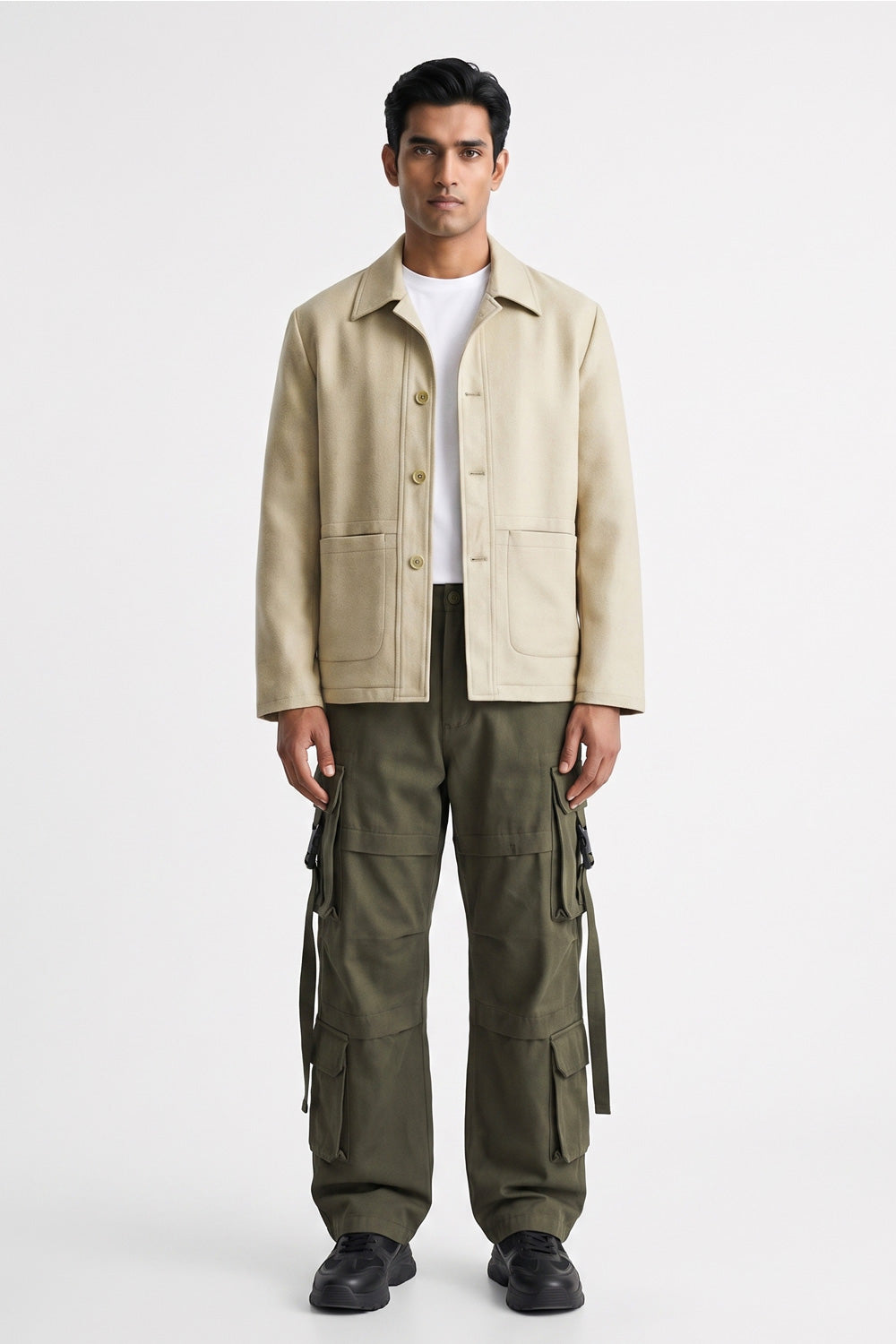 A man stands against a light gray studio background wearing a beige utility linen jacket over a white T-shirt, olive green cargo pants, and black shoes.