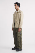 A man wearing a light beige linen overshirt, olive green cargo pants, and black shoes, standing in profile against a plain light gray studio background.
