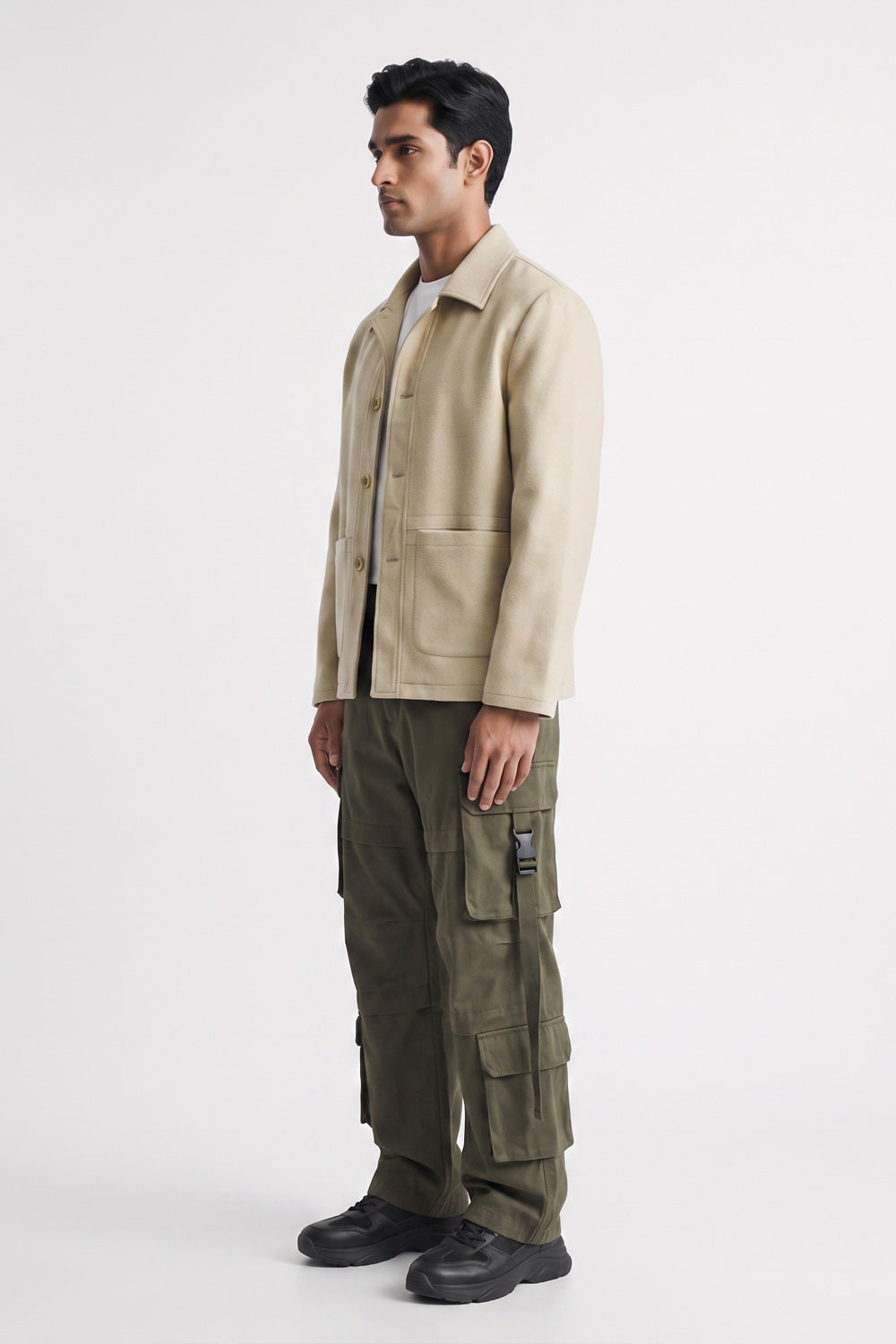 A man wearing a light beige linen overshirt, olive green cargo pants, and black shoes, standing in profile against a plain light gray studio background.