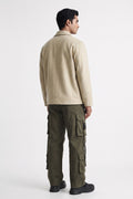 A man shown from the back wearing a beige long-sleeve linen overshirt and olive green cargo pants against a plain light gray studio background.