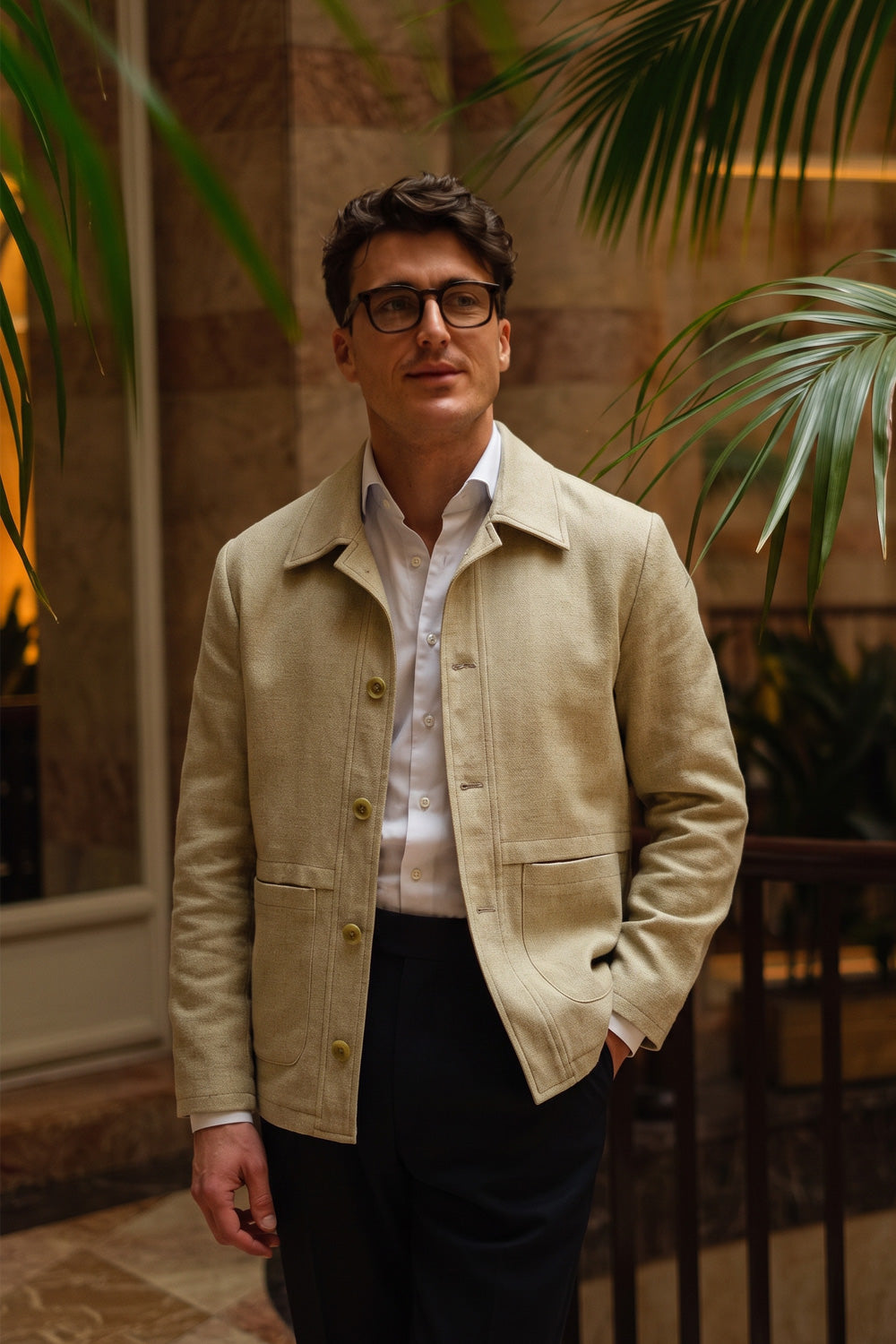 A man wearing a light beige clinen jacket over a white button-down shirt and dark trousers, standing in an indoor setting with warm lighting and palm leaves in the background.