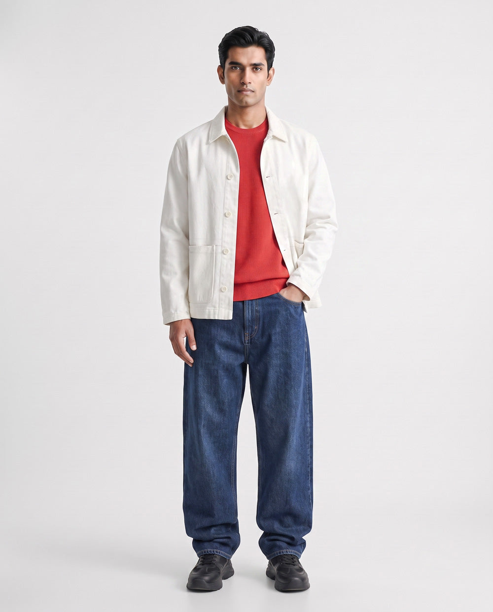 A man modeling a white button-up linen overshirt over a red crewneck sweater, paired with loose blue jeans and black shoes, against a plain light gray studio background.