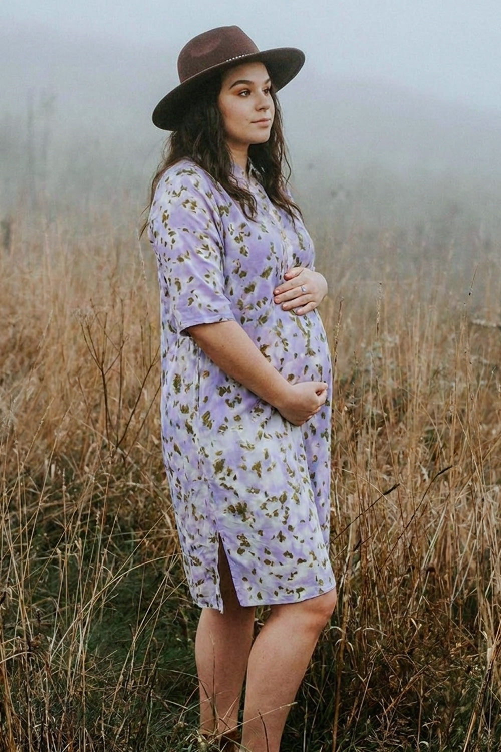 A pregnant woman wearing a light purple floral eco-printed maternity tunic dress and a brown wide-brim hat, standing in a misty field of dry grass.