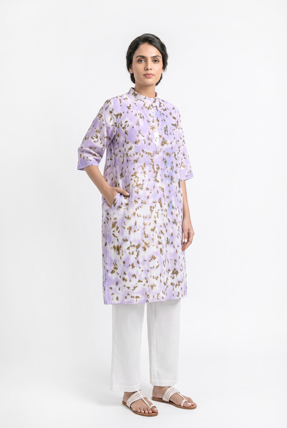 A model wearing a lavender cotton eco-print kurta featuring natural marigold leaf motifs in earthy golden tones. The straight-fit kurta has a mandarin collar, three-quarter sleeves, a short button placket, and side pockets. It is paired with off-white straight pants and white braided sandals against a clean white background.