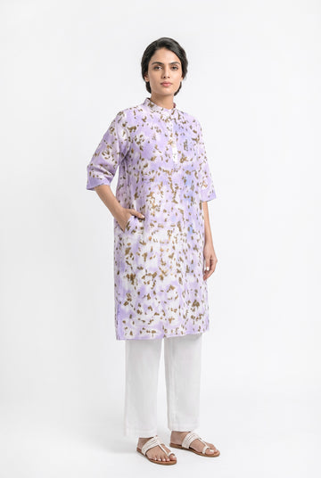 A model wearing a lavender cotton eco-print kurta featuring natural marigold leaf motifs in earthy golden tones. The straight-fit kurta has a mandarin collar, three-quarter sleeves, a short button placket, and side pockets. It is paired with off-white straight pants and white braided sandals against a clean white background.