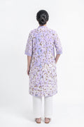 A woman viewed from the back wearing a lavender purple tunic with a brown and gold floral pattern, paired with white pants, against a plain white studio background.