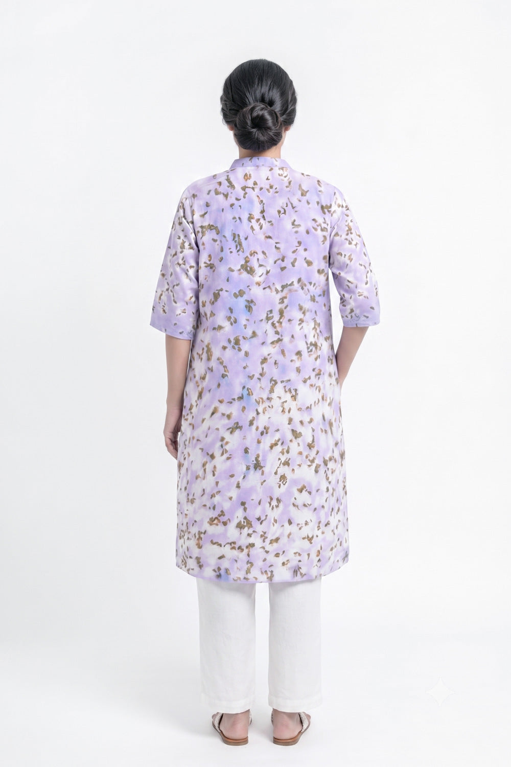 A woman viewed from the back wearing a lavender purple tunic with a brown and gold floral pattern, paired with white pants, against a plain white studio background.