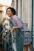 A woman wearing a lavender floral dress leans against a balcony railing on a sunlit street, smiling and looking upward.