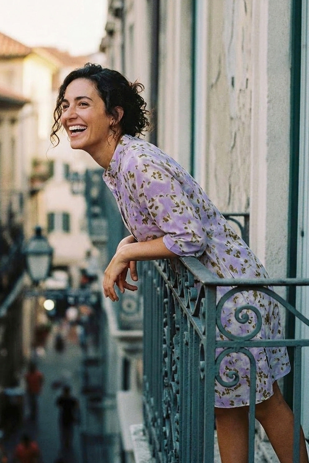 A woman wearing a lavender floral dress leans against a balcony railing on a sunlit street, smiling and looking upward.