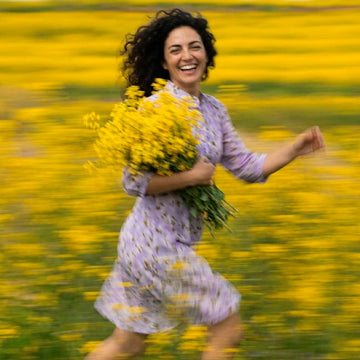 A woman with curly dark hair runs through a field of yellow flowers while holding a large bouquet of yellow flowers. She is wearing a light purple patterned dress.