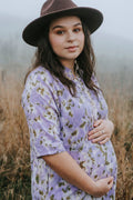 A pregnant woman wearing a lavender short-sleeve button-up shirt with a green floral botanical-print pattern and a brown wide-brim hat, standing in a misty field.