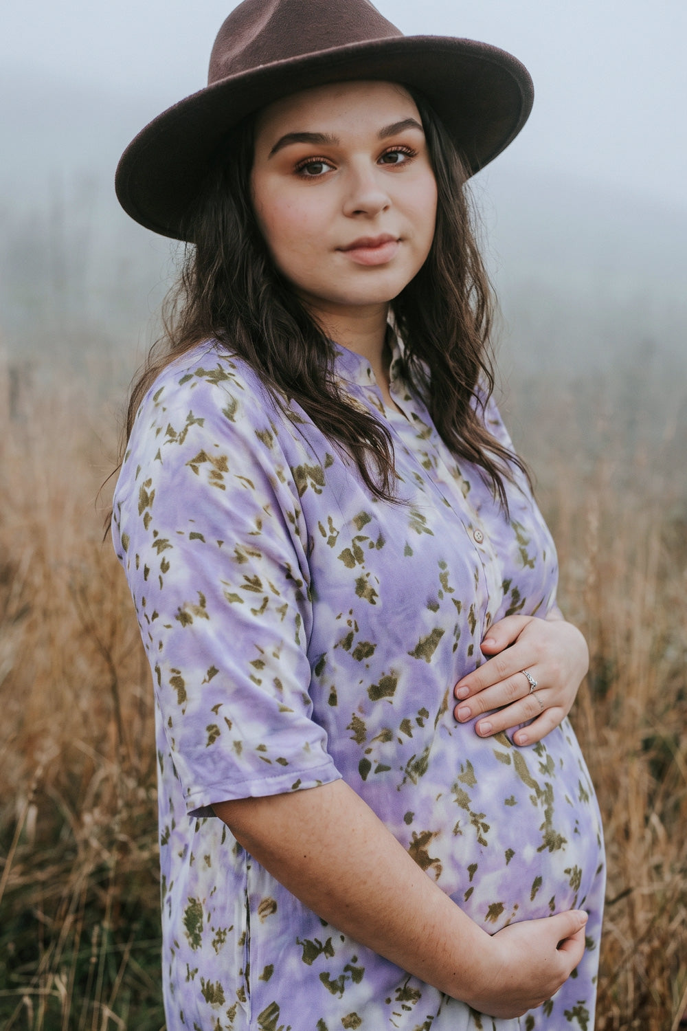 A pregnant woman wearing a lavender short-sleeve button-up shirt with a green floral botanical-print pattern and a brown wide-brim hat, standing in a misty field.