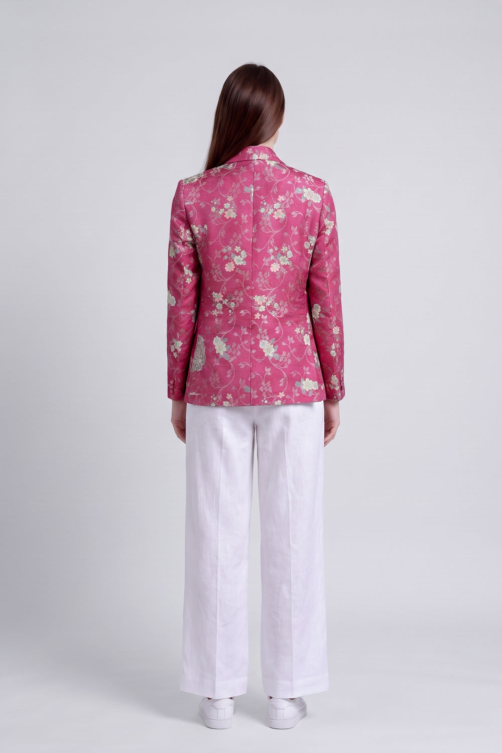 A model shown from the back wearing a pink floral silk blazer and white wide-leg trousers against a light gray studio background.