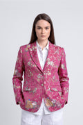 A woman modeling a pink floral silk jacquard blazer with detailed embroidery on the front pockets, worn over a white collared shirt and white trousers, against a light gray studio background.