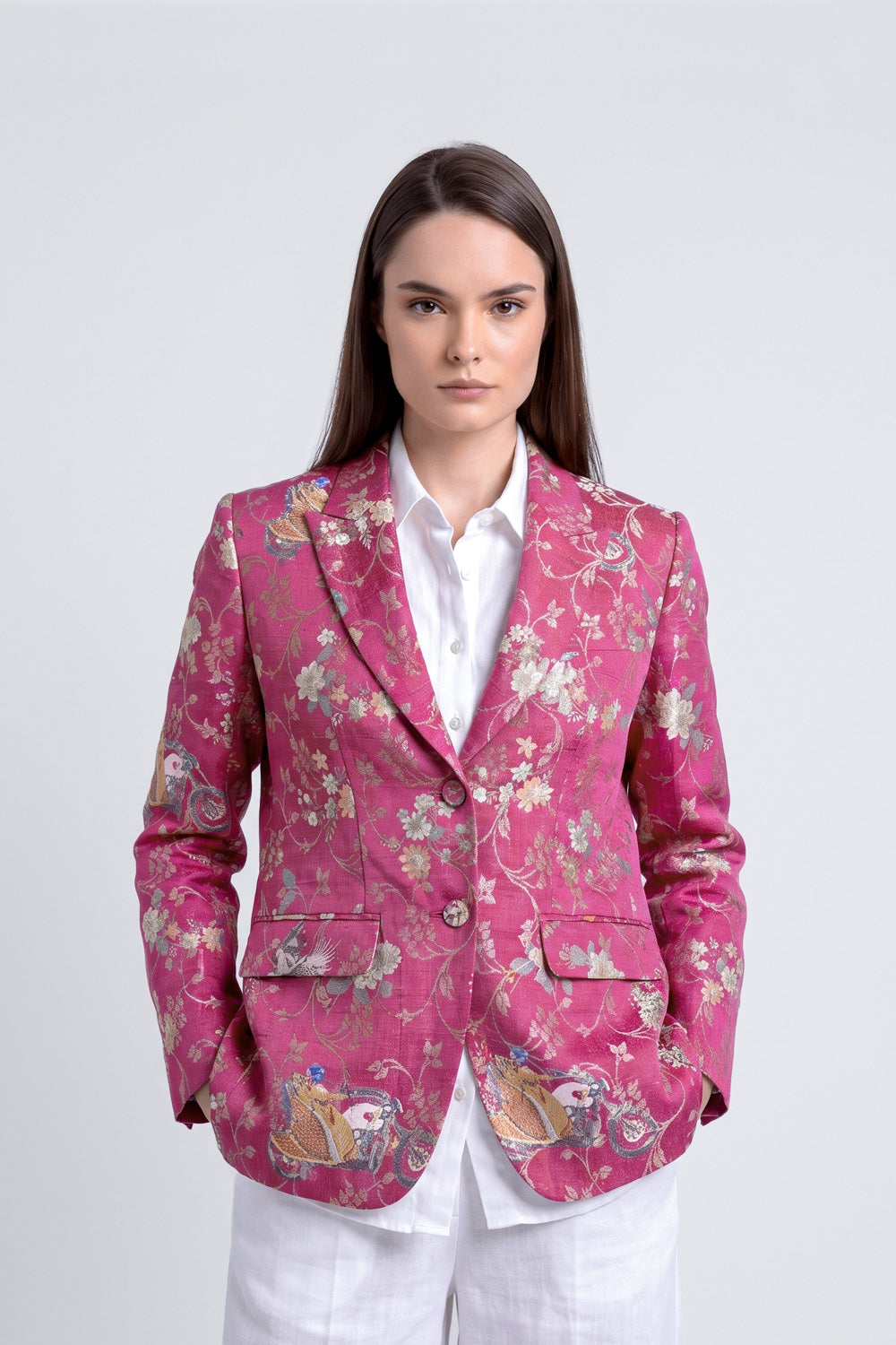A woman modeling a pink floral silk jacquard blazer with detailed embroidery on the front pockets, worn over a white collared shirt and white trousers, against a light gray studio background.