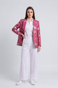 A model stands against a light gray studio background wearing a pink floral silk blazer over a white button-up shirt and white wide-leg trousers, with white sneakers.