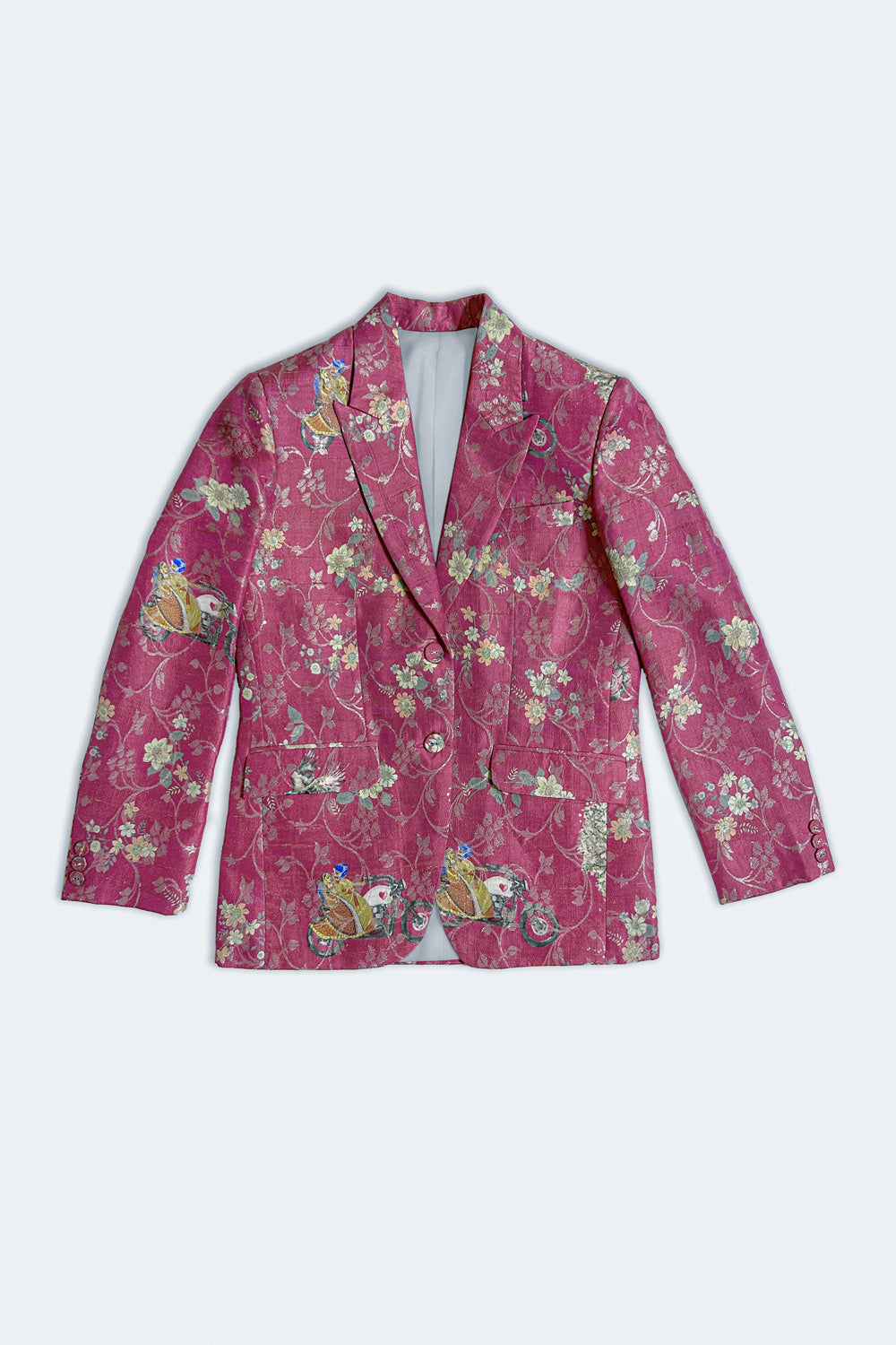 A model stands against a light gray studio background wearing a white button-up shirt and white wide-leg trousers, with a shiny magenta floral silk blazer draped over her shoulders and white sneakers.