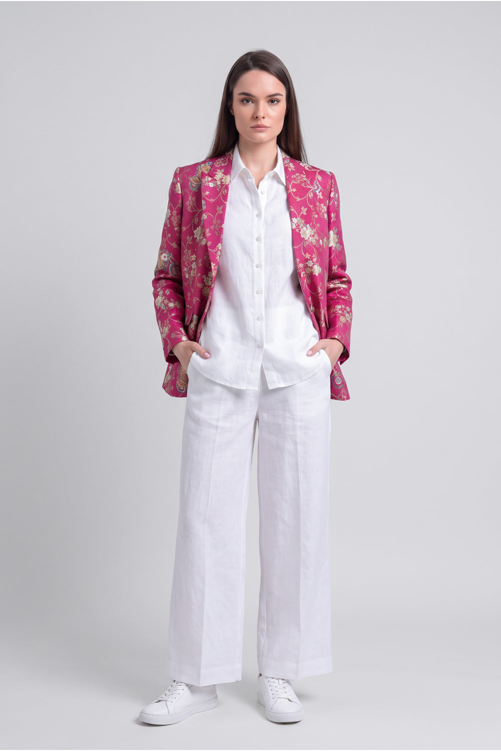 A model stands against a light gray studio background wearing a white button-up shirt and white wide-leg trousers, with a shiny magenta floral silk blazer draped over her shoulders and white sneakers.