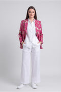 A model stands against a light gray studio background wearing a white button-up shirt and white wide-leg trousers, with a shiny magenta floral silk blazer draped over her shoulders and white sneakers.