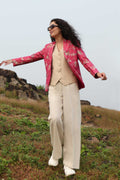 A model stands against a green outdoor background wearing a beige button-up vest and white wide-leg trousers, with a shiny magenta floral silk blazer draped over her shoulders and white sneakers.