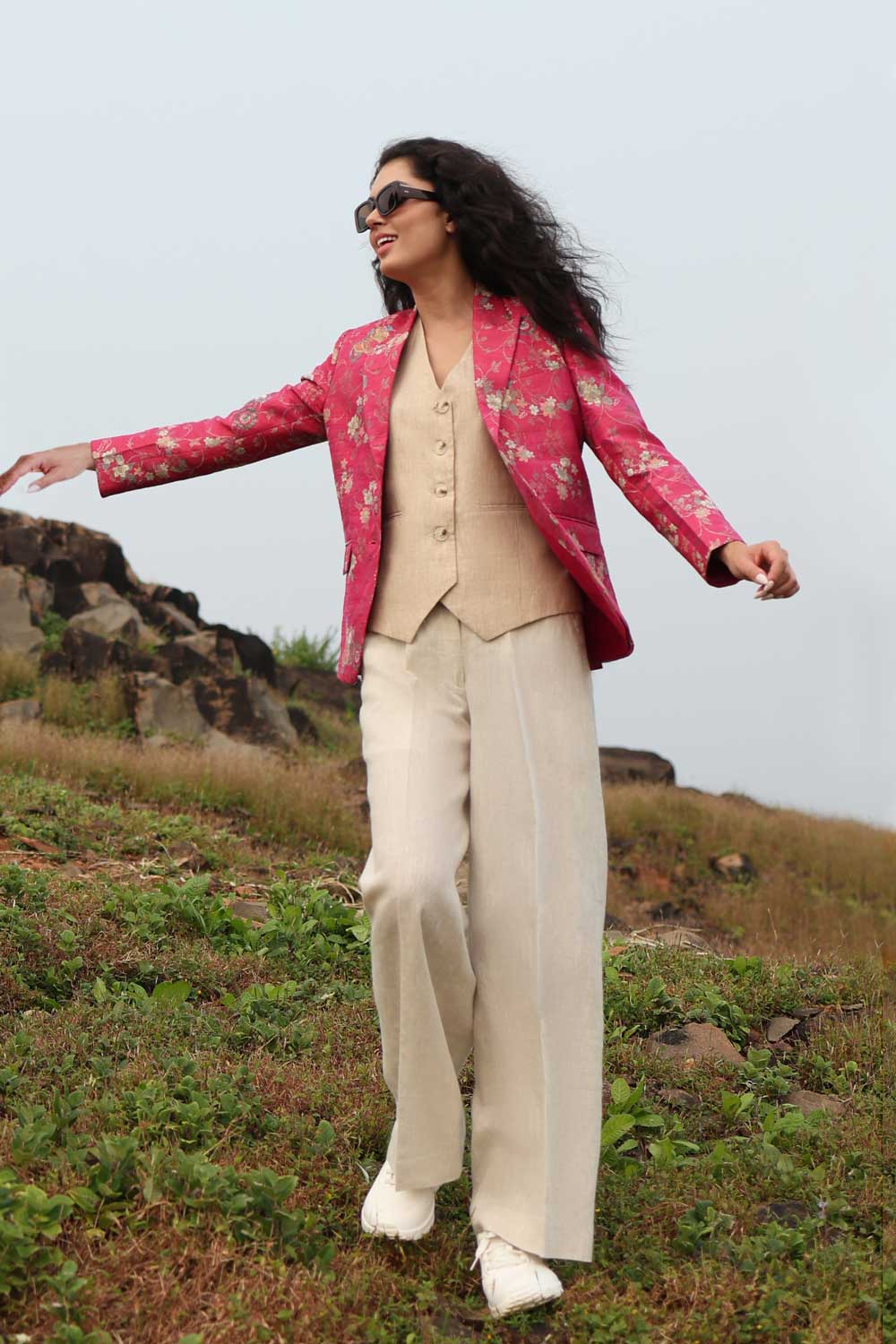 A model stands against a green outdoor background wearing a beige button-up vest and white wide-leg trousers, with a shiny magenta floral silk blazer draped over her shoulders and white sneakers.