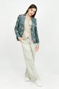 A model stands against a white studio background wearing a beige button-up shirt and white wide-leg trousers, with a shiny teal floral silk blazer draped over her shoulders and white sneakers.