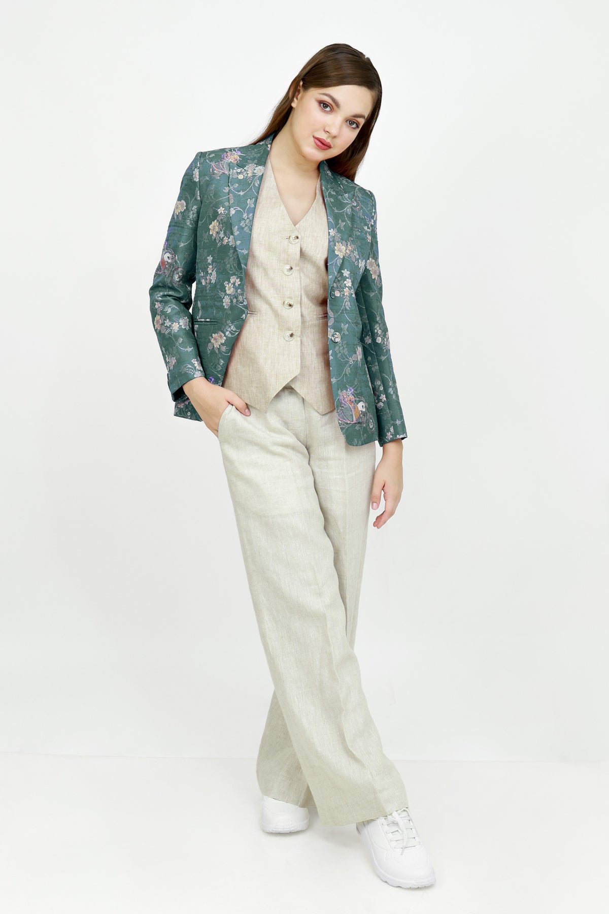 A model stands against a white studio background wearing a beige button-up shirt and white wide-leg trousers, with a shiny teal floral silk blazer draped over her shoulders and white sneakers.