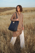 A woman standing in a dry grassy field holds a large dark navy tote bag. She is wearing a brown linen shirt and white linen pants, with rolling hills in the background under a soft sky.