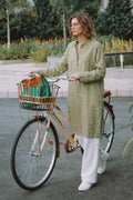 A woman wearing a sage green linen tunic and white wide-leg trousers stands next to a vintage wooden bicycle with a green and brown bag in its basket, set against a blurred outdoor garden background.