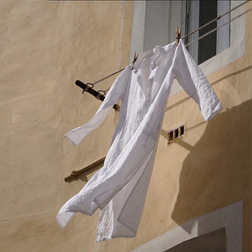 A white long-sleeve linen shirt hangs on a clothesline with wooden clothespins against a textured beige stucco wall.