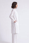 A woman shown in profile wearing a loose white long-sleeve linen tunic top and matching white pants against a plain light gray studio background.