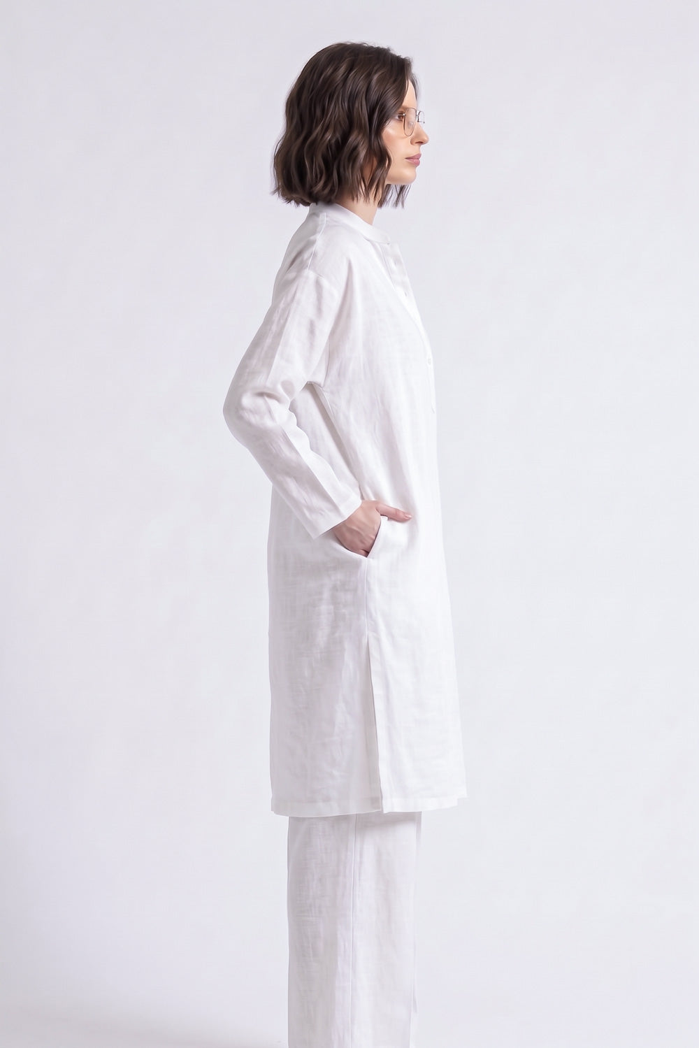 A woman shown in profile wearing a loose white long-sleeve linen tunic top and matching white pants against a plain light gray studio background.