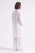 A model shown from the back wearing a long white linen tunic and matching wide-leg trousers against a light lavender studio background.