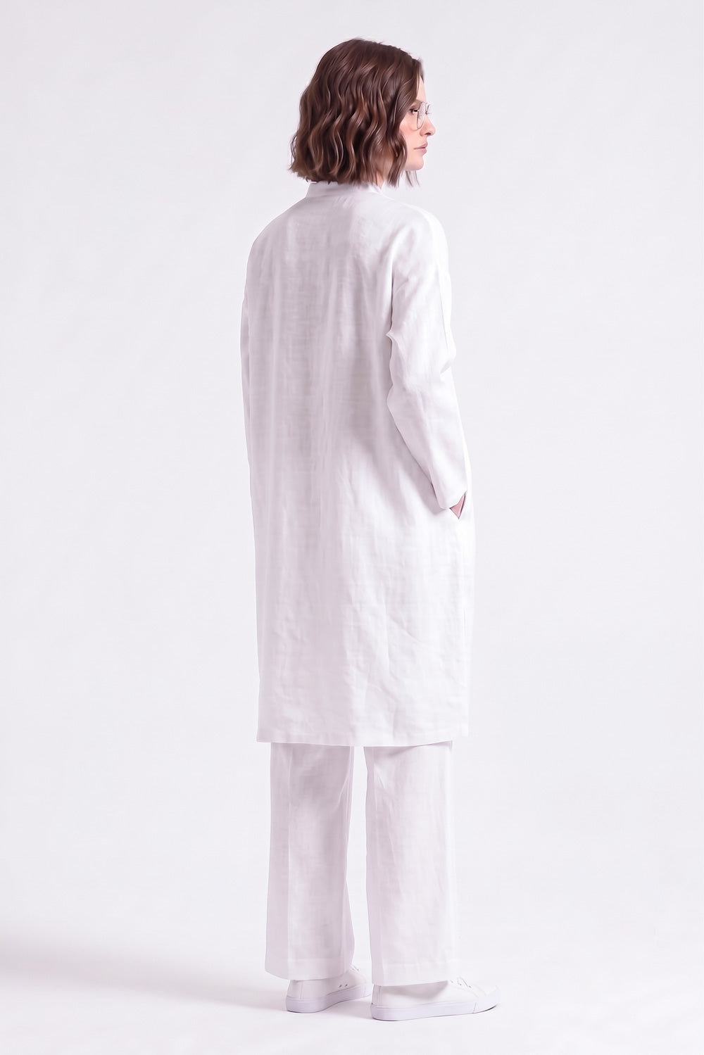 A model shown from the back wearing a long white linen tunic and matching wide-leg trousers against a light lavender studio background.
