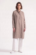 A woman modeling a long-sleeved, knee-length taupe linen tunic with a mandarin collar and button placket, paired with white trousers and white sneakers, against a plain white studio background.