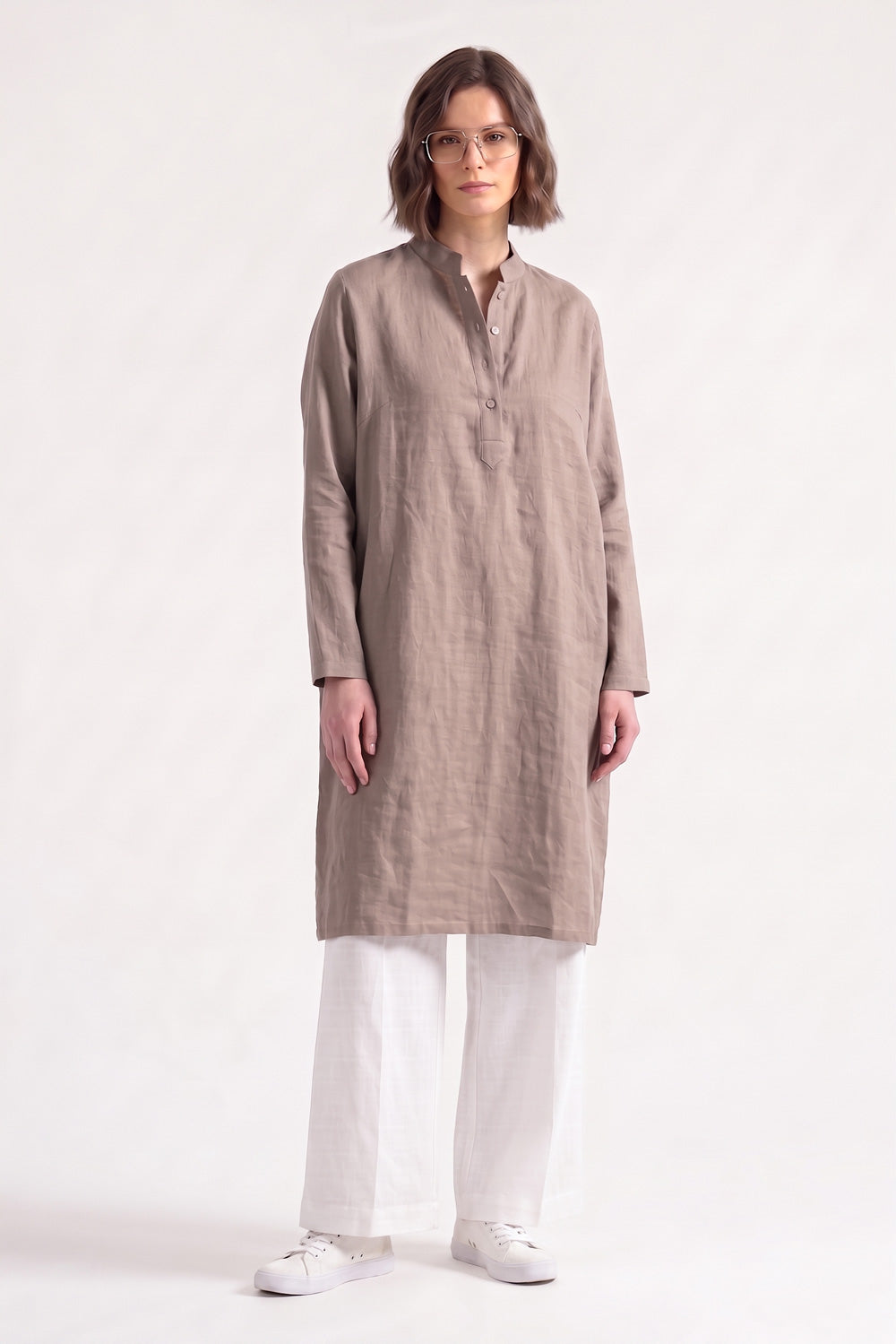A woman modeling a long-sleeved, knee-length taupe linen tunic with a mandarin collar and button placket, paired with white trousers and white sneakers, against a plain white studio background.