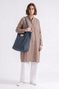 A model wearing a beige linen tunic, white pants, and white sneakers is shown from the waist up, carrying a large navy blue canvas tote bag with a shoulder strap. The background is a clean, off-white studio backdrop.