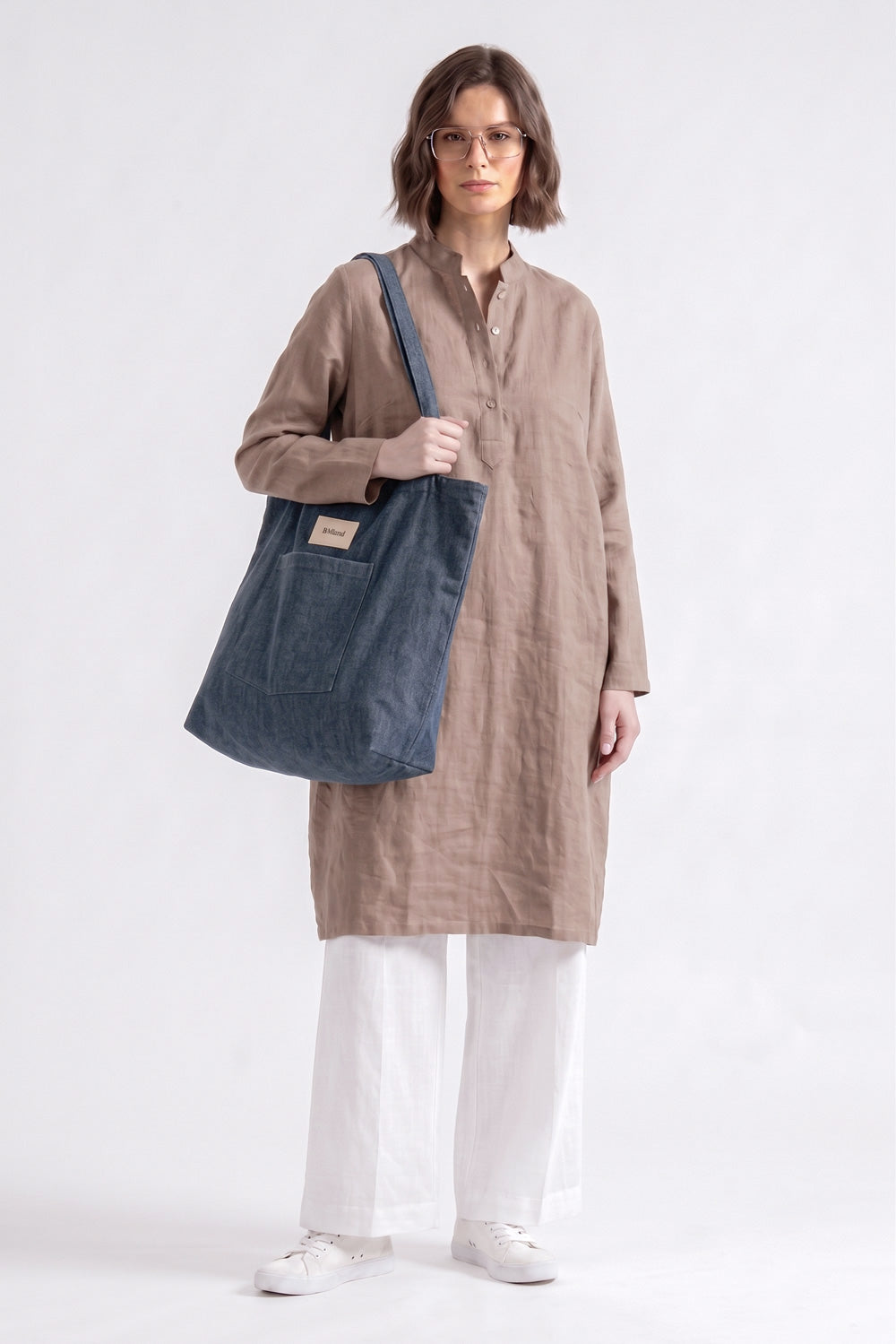 A model wearing a beige linen tunic, white pants, and white sneakers is shown from the waist up, carrying a large navy blue canvas tote bag with a shoulder strap. The background is a clean, off-white studio backdrop.