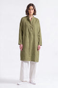 A woman modeling a long-sleeved olive green linen tunic with a mandarin collar and button placket, paired with white wide-leg trousers and white sneakers, against a plain light pink studio background.