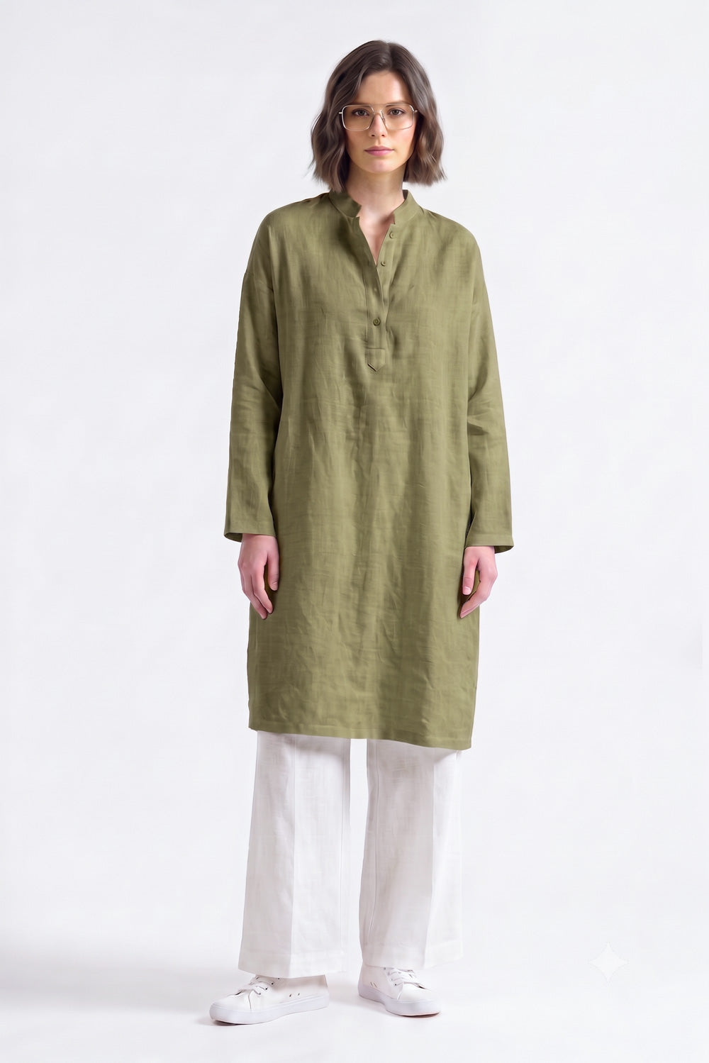 A woman modeling a long-sleeved olive green linen tunic with a mandarin collar and button placket, paired with white wide-leg trousers and white sneakers, against a plain light pink studio background.
