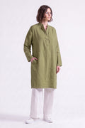 A woman modeling a long-sleeved olive green linen tunic with a split neckline and button placket, paired with white wide-leg trousers and white sneakers, set against a solid pale pink studio background.