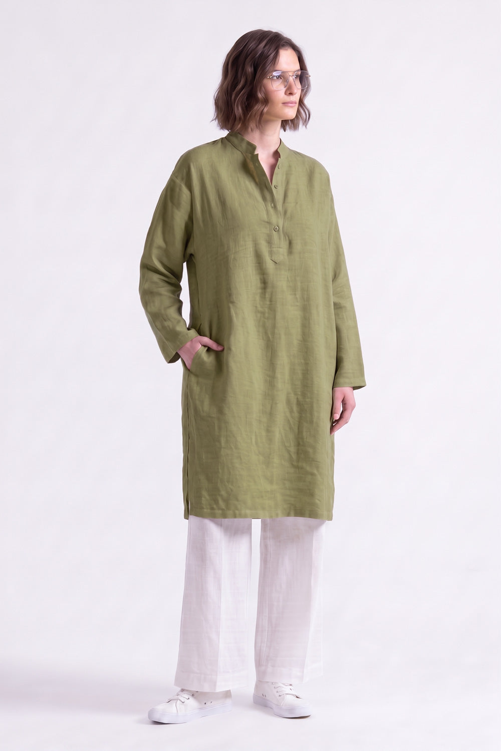 A woman modeling a long-sleeved olive green linen tunic with a split neckline and button placket, paired with white wide-leg trousers and white sneakers, set against a solid pale pink studio background.